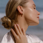 A blonde woman in a low bun smiles softly with closed eyes by the beach, wearing a light satin top, gold hoop earrings, and the LLUME Jewelry Vacation Capsule Set—a versatile necklace set perfect for stacking.