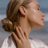 A blonde woman in a low bun smiles softly with closed eyes by the beach, wearing a light satin top, gold hoop earrings, and the LLUME Jewelry Vacation Capsule Set—a versatile necklace set perfect for stacking.