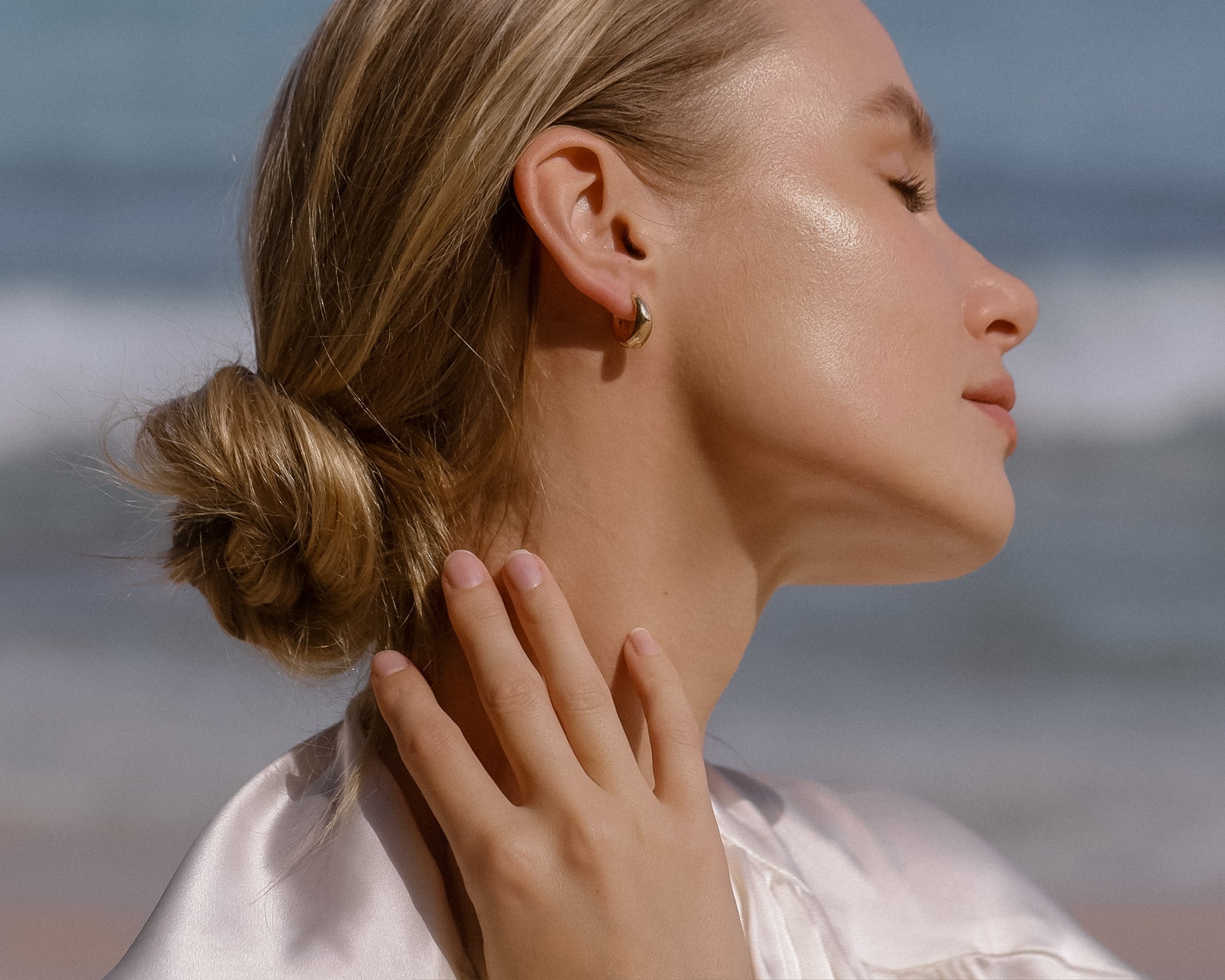 A blonde woman in a low bun smiles softly with closed eyes by the beach, wearing a light satin top, gold hoop earrings, and the LLUME Jewelry Vacation Capsule Set—a versatile necklace set perfect for stacking.
