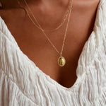 A person styled in a textured white blouse and LLUME Jewelry's Vintage Daily Set—layered gold chains with an elegant oval pendant—stands against a warm-toned background.