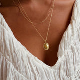 A person styled in a textured white blouse and LLUME Jewelry's Vintage Daily Set—layered gold chains with an elegant oval pendant—stands against a warm-toned background.