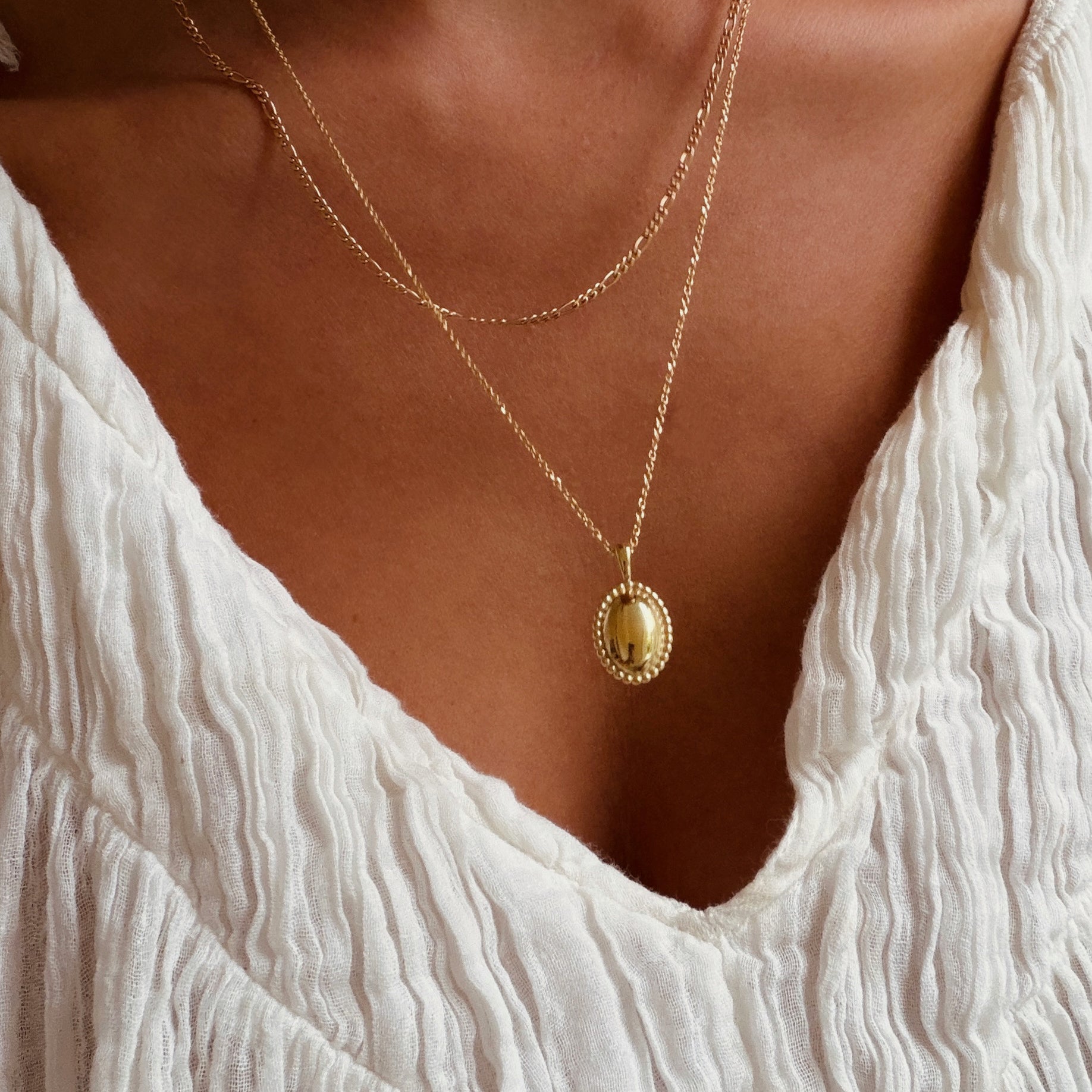 A person styled in a textured white blouse and LLUME Jewelry's Vintage Daily Set—layered gold chains with an elegant oval pendant—stands against a warm-toned background.