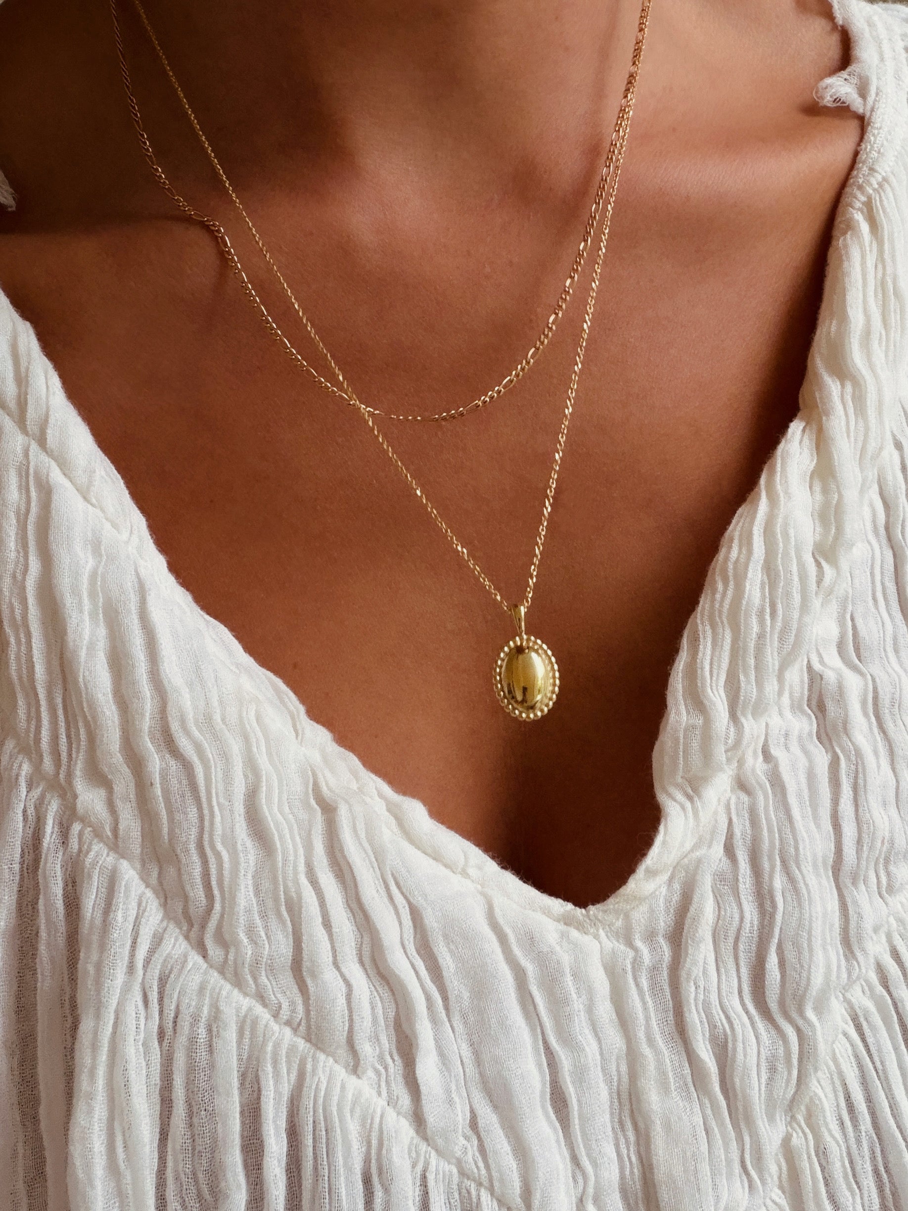 A person styled in a textured white blouse and LLUME Jewelry's Vintage Daily Set—layered gold chains with an elegant oval pendant—stands against a warm-toned background.