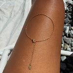 The LLUME Jewelry Amber Anklet—a delicate gold piece adorned with a dangling charm—rests gracefully on a sun-kissed leg, radiating everyday elegance against a backdrop of pebbles and a white towel.