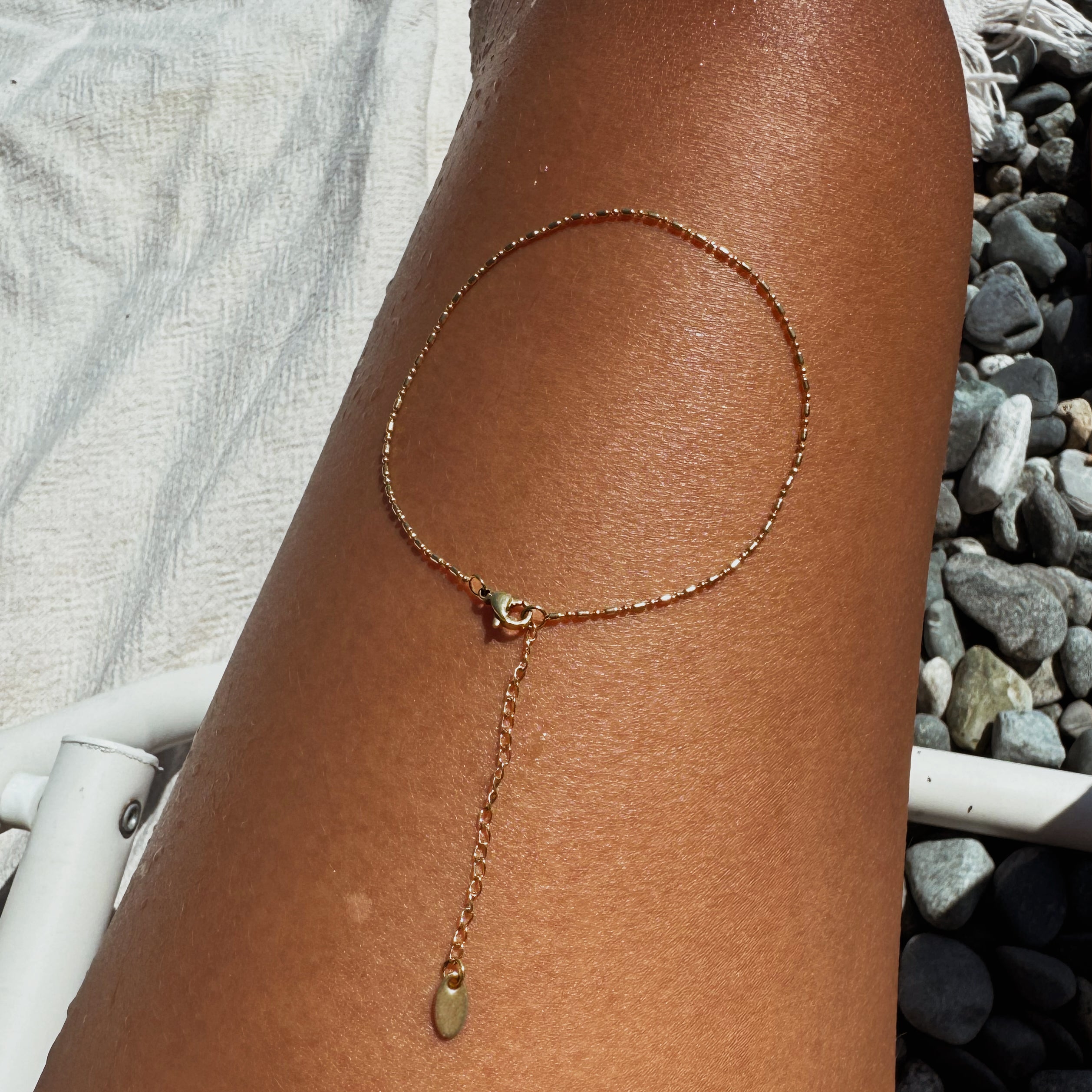 The LLUME Jewelry Amber Anklet—a delicate gold piece adorned with a dangling charm—rests gracefully on a sun-kissed leg, radiating everyday elegance against a backdrop of pebbles and a white towel.