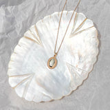 The Aurelia Vintage Oval Necklace by LLUME Jewelry, featuring a white stone pendant, is displayed on a shiny iridescent shell-shaped dish atop crumpled white paper.