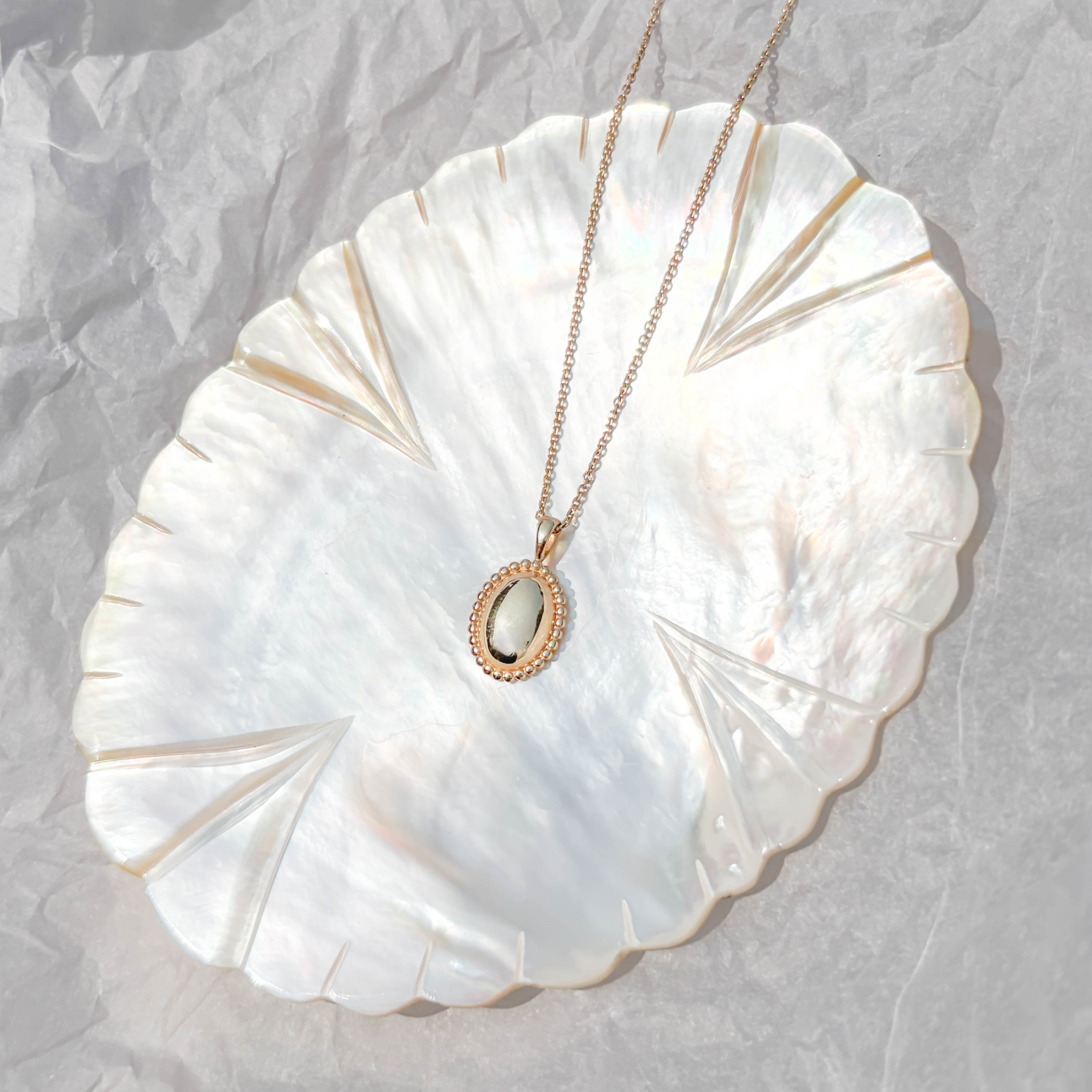 The Aurelia Vintage Oval Necklace by LLUME Jewelry, featuring a white stone pendant, is displayed on a shiny iridescent shell-shaped dish atop crumpled white paper.
