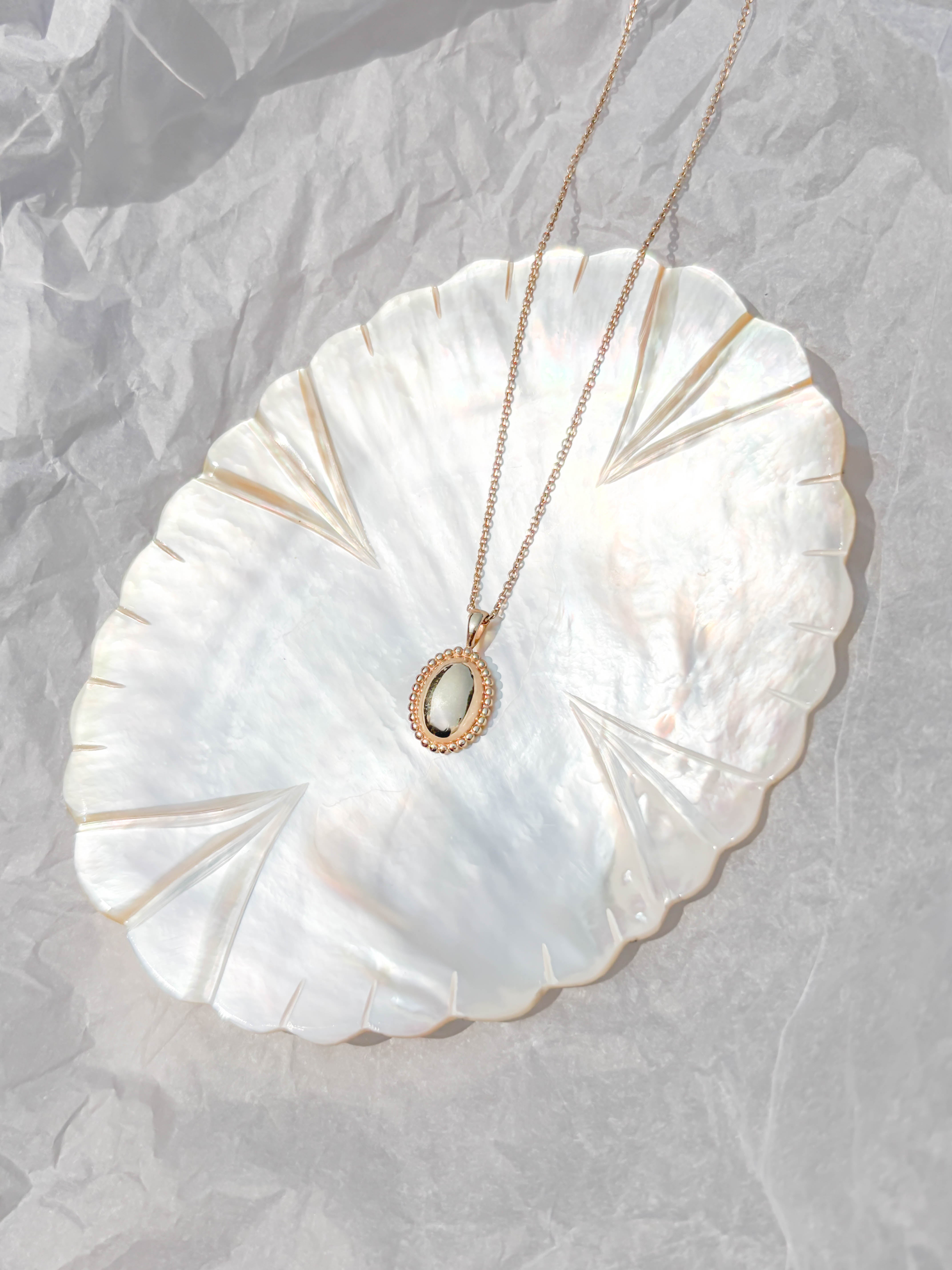 The Aurelia Vintage Oval Necklace by LLUME Jewelry, featuring a white stone pendant, is displayed on a shiny iridescent shell-shaped dish atop crumpled white paper.