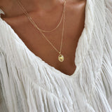 A person wearing a white, textured, V-neck blouse and the Vintage Daily Set by LLUME Jewelry—a layered necklace set with two delicate gold chains, featuring an elegant pendant and a small vintage oval necklace—shown from neck to upper chest.