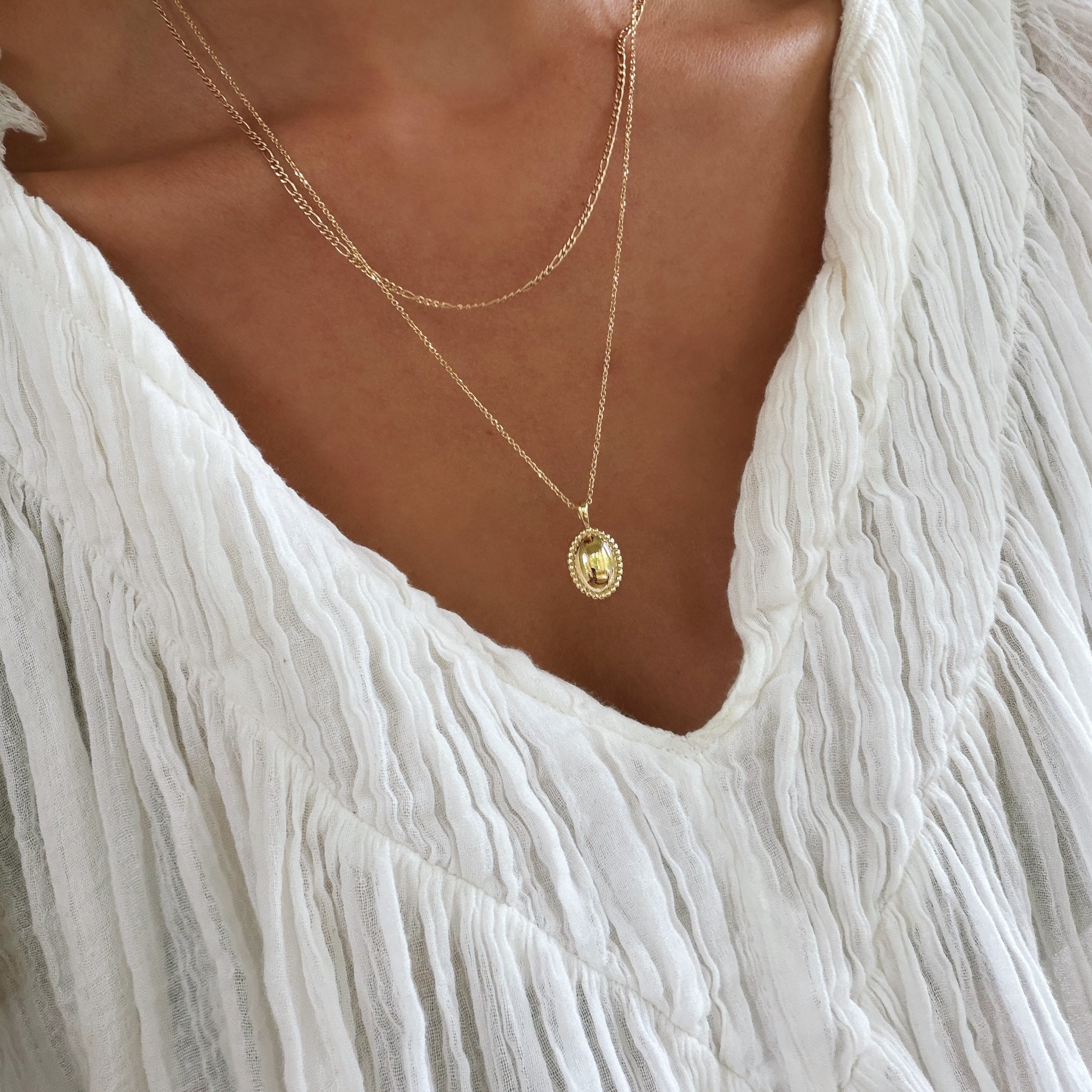 A person wearing a white, textured, V-neck blouse and the Vintage Daily Set by LLUME Jewelry—a layered necklace set with two delicate gold chains, featuring an elegant pendant and a small vintage oval necklace—shown from neck to upper chest.