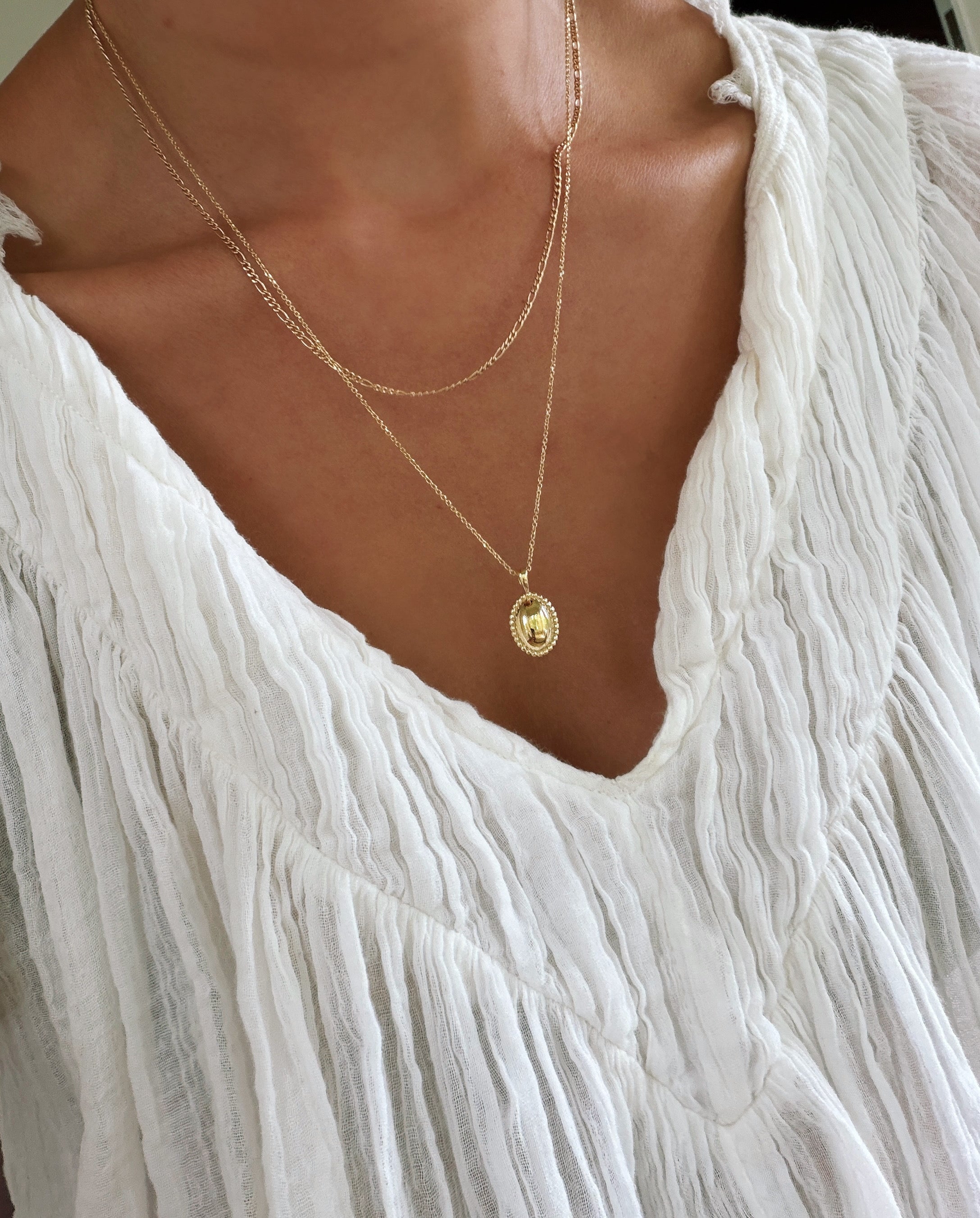 A person wearing a white, textured, V-neck blouse and the Vintage Daily Set by LLUME Jewelry—a layered necklace set with two delicate gold chains, featuring an elegant pendant and a small vintage oval necklace—shown from neck to upper chest.