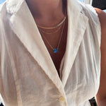 Wearing LLUME Jewelry’s Seacove Favorites Set—a combination of layered gold necklaces featuring the skinny hailey chain and a delicate seacove necklace with a blue rectangular pendant—a person in a sleeveless white collared blouse stands by a window, bathed in bright sunlight.