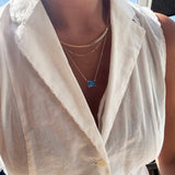Wearing LLUME Jewelry’s Seacove Favorites Set—a combination of layered gold necklaces featuring the skinny hailey chain and a delicate seacove necklace with a blue rectangular pendant—a person in a sleeveless white collared blouse stands by a window, bathed in bright sunlight.