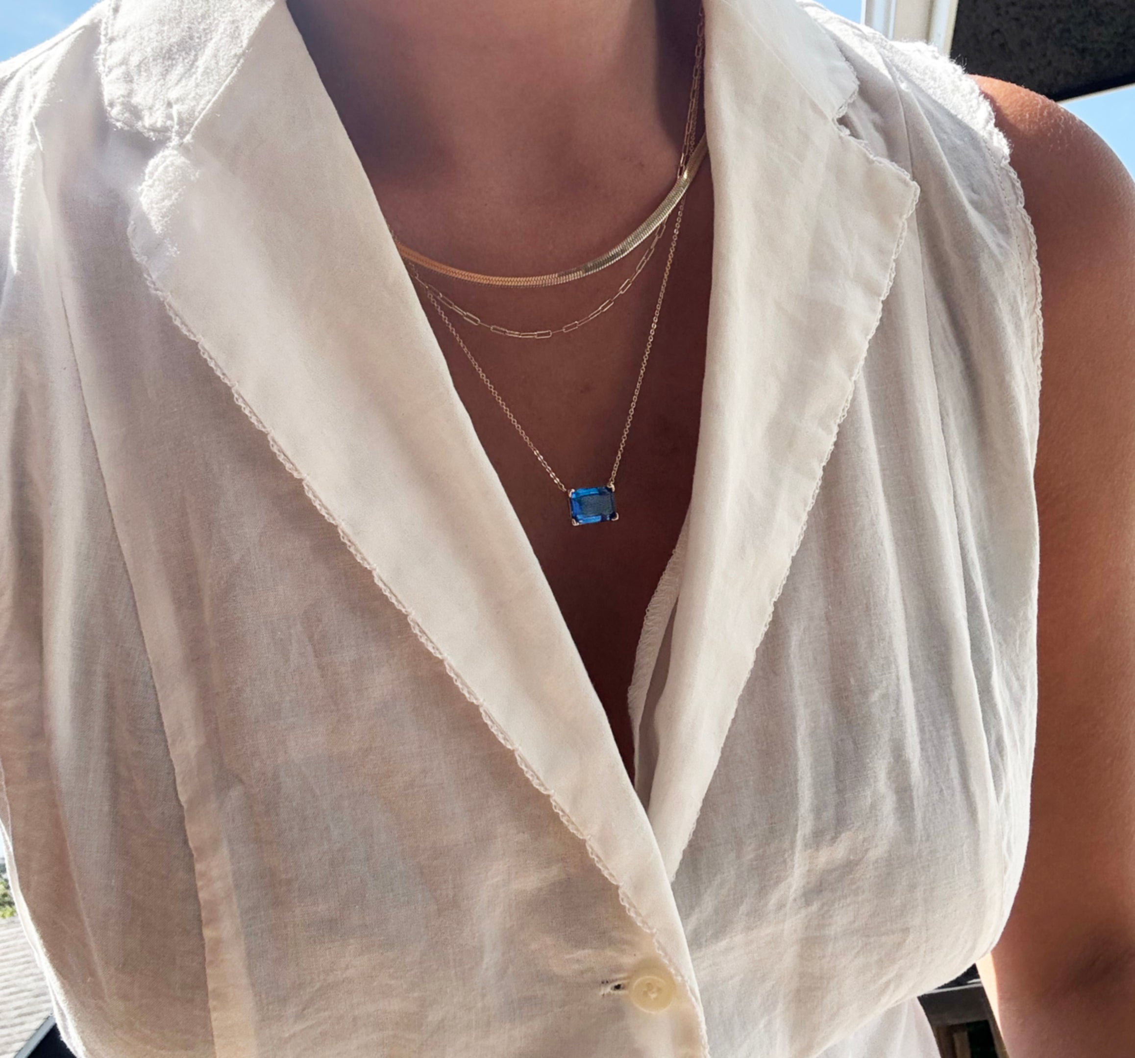 Wearing LLUME Jewelry’s Seacove Favorites Set—a combination of layered gold necklaces featuring the skinny hailey chain and a delicate seacove necklace with a blue rectangular pendant—a person in a sleeveless white collared blouse stands by a window, bathed in bright sunlight.