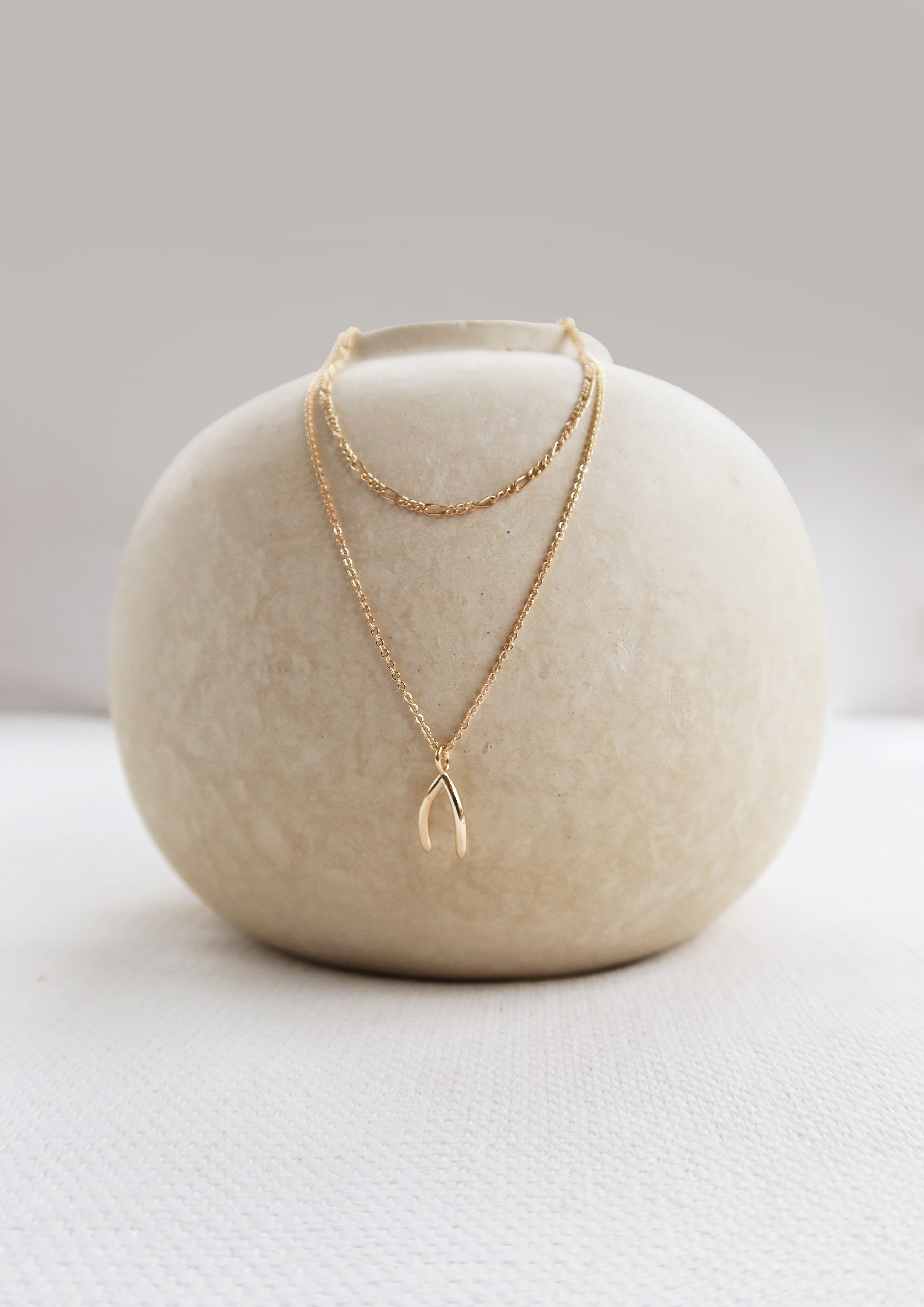 A round, off-white ceramic vase displays the LLUME Jewelry Wishbone Daily Set—featuring the Colette Necklace and Wishbone Necklace—elegantly arranged against a neutral background.