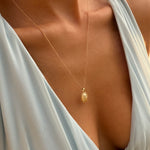 A close-up of a person wearing a pale blue, deep V-neck dress and the LLUME Jewelry Aurelia Vintage Oval Necklace, with warm sunlight highlighting their skin.