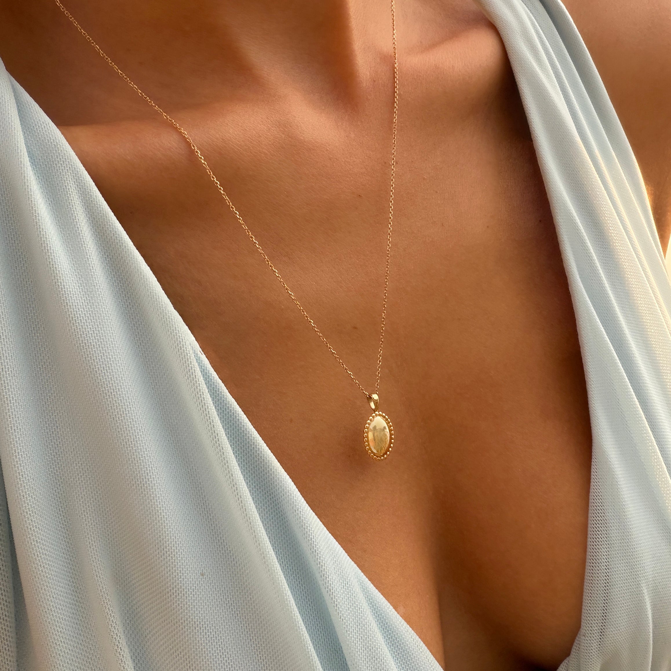 A close-up of a person wearing a pale blue, deep V-neck dress and the LLUME Jewelry Aurelia Vintage Oval Necklace, with warm sunlight highlighting their skin.