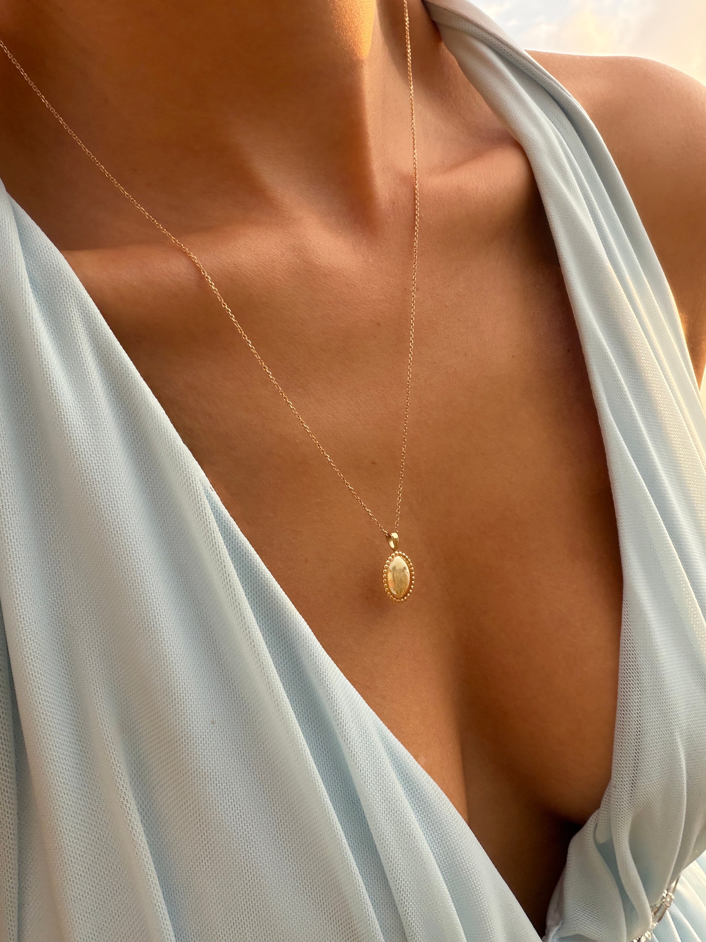 A close-up of a person wearing a pale blue, deep V-neck dress and the LLUME Jewelry Aurelia Vintage Oval Necklace, with warm sunlight highlighting their skin.