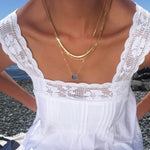 Wearing the Seacove Favorites Set by LLUME Jewelry, which includes layered gold necklaces—a slender hailey chain and one with a blue pendant—a person in a white lace-trimmed dress sits on a rocky beach with water in the background.