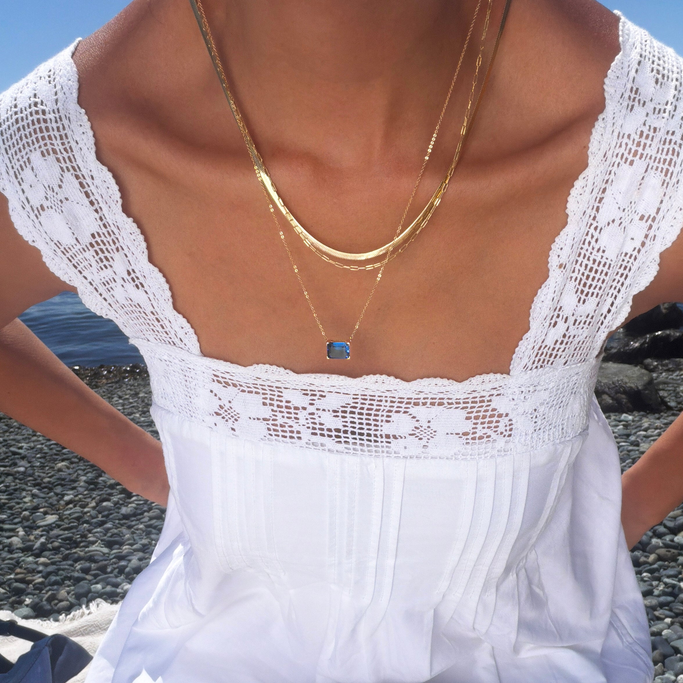 Wearing the Seacove Favorites Set by LLUME Jewelry, which includes layered gold necklaces—a slender hailey chain and one with a blue pendant—a person in a white lace-trimmed dress sits on a rocky beach with water in the background.