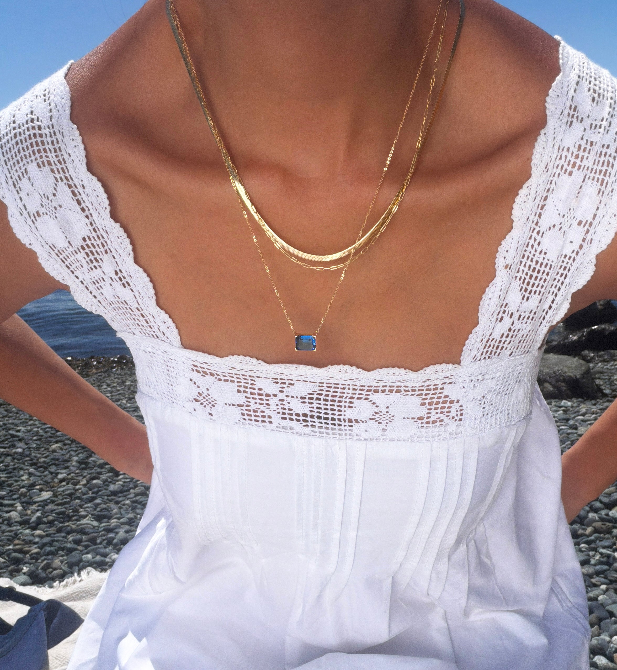 Wearing the Seacove Favorites Set by LLUME Jewelry, which includes layered gold necklaces—a slender hailey chain and one with a blue pendant—a person in a white lace-trimmed dress sits on a rocky beach with water in the background.