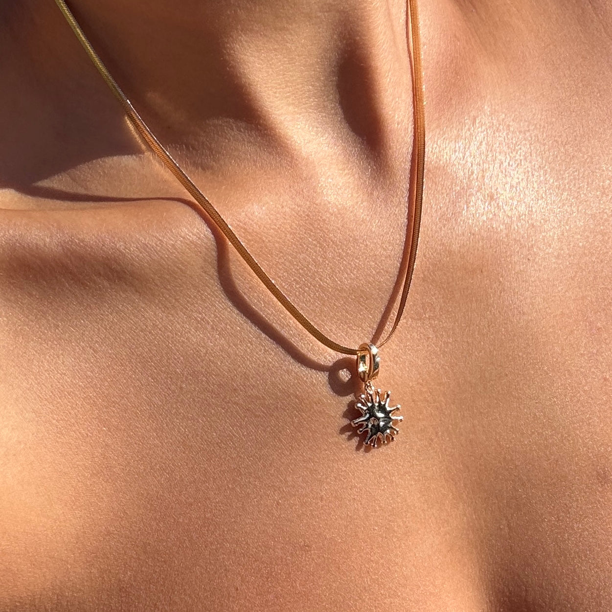 A close-up of LLUME Jewelry's Every Occasion Set shows a gold sun-shaped charm necklace with a snake chain resting on bare skin, its warm shine beautifully highlighted by the sunlight.