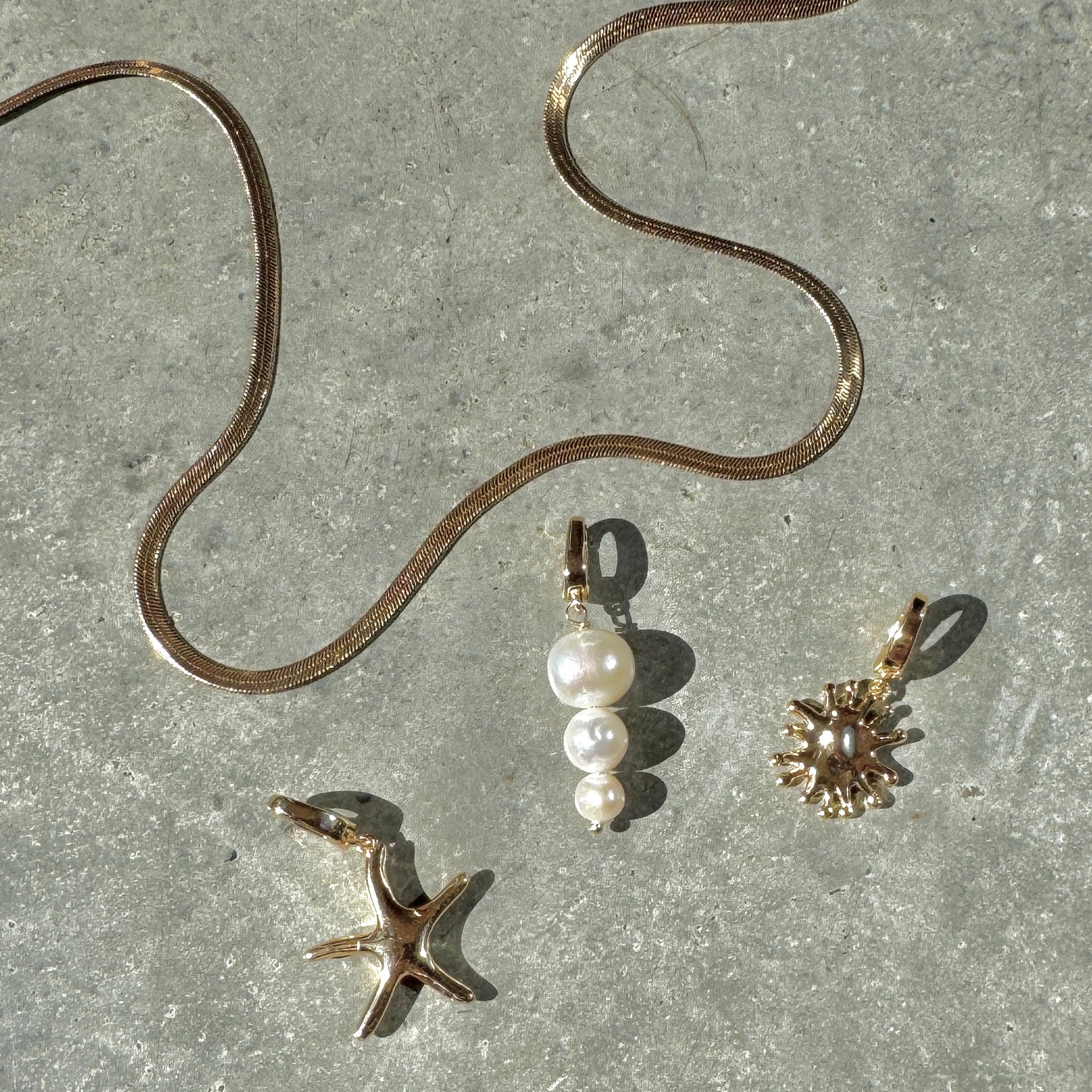 The LLUME Jewelry Every Occasion Set features a gold snake chain necklace on a gray textured surface, accompanied by three interchangeable charms: a starfish, white pearl cluster, and sunburst design.