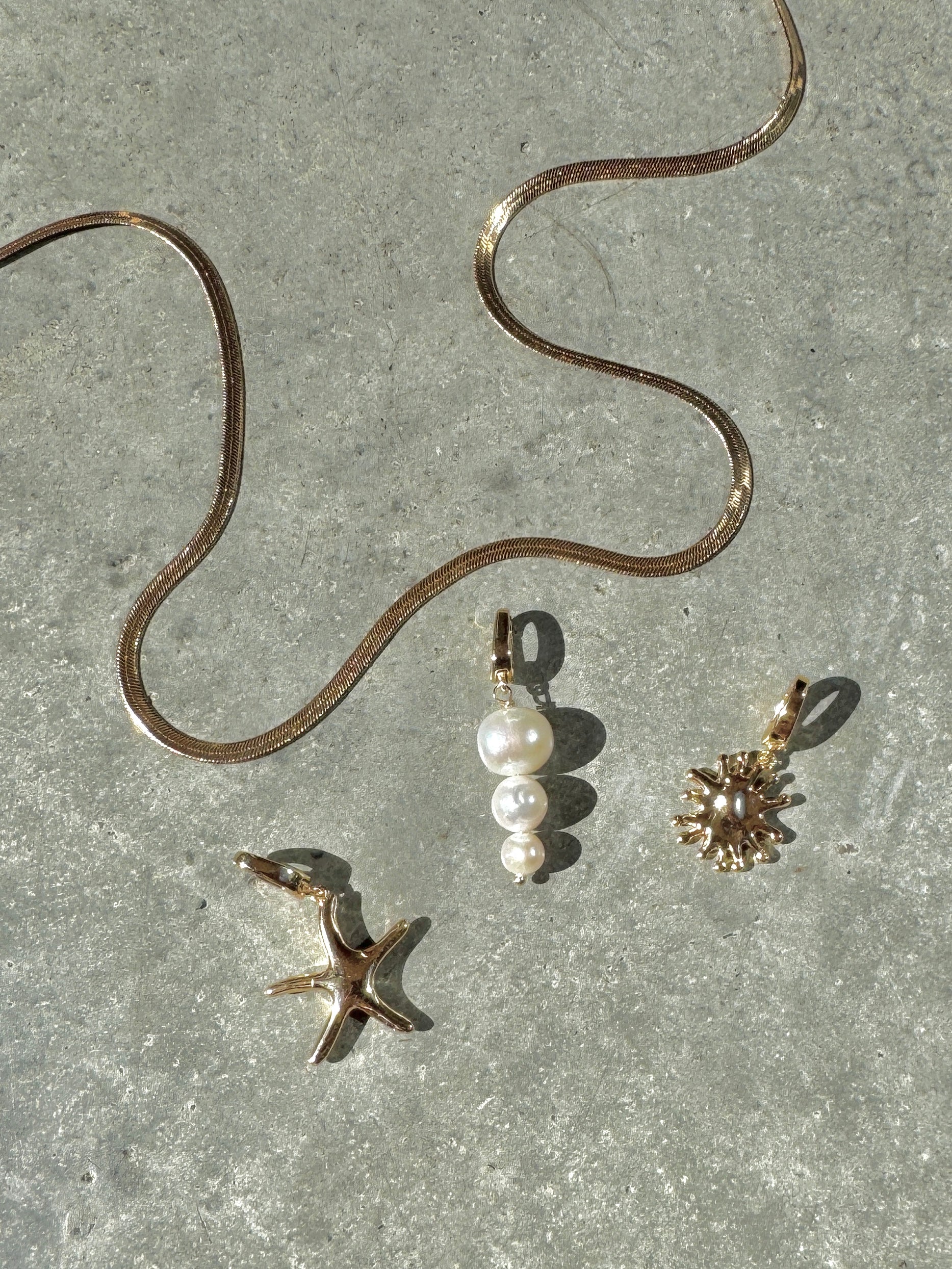 The LLUME Jewelry Every Occasion Set features a gold snake chain necklace on a gray textured surface, accompanied by three interchangeable charms: a starfish, white pearl cluster, and sunburst design.