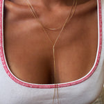 A person wearing a white tank top with red trim is shown layered in elegant jewelry, including the Dusk Necklace by LLUME Jewelry and a gold lariat necklace. The photo highlights their collarbone, chest, and the featured jewelry.