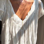 A person wearing a textured white blouse with a deep V-neckline, elegantly adorned with the LLUME Jewelry Dusk Necklace—a refined gold lariat piece—layered alongside other delicate necklaces and softly illuminated by natural sunlight.