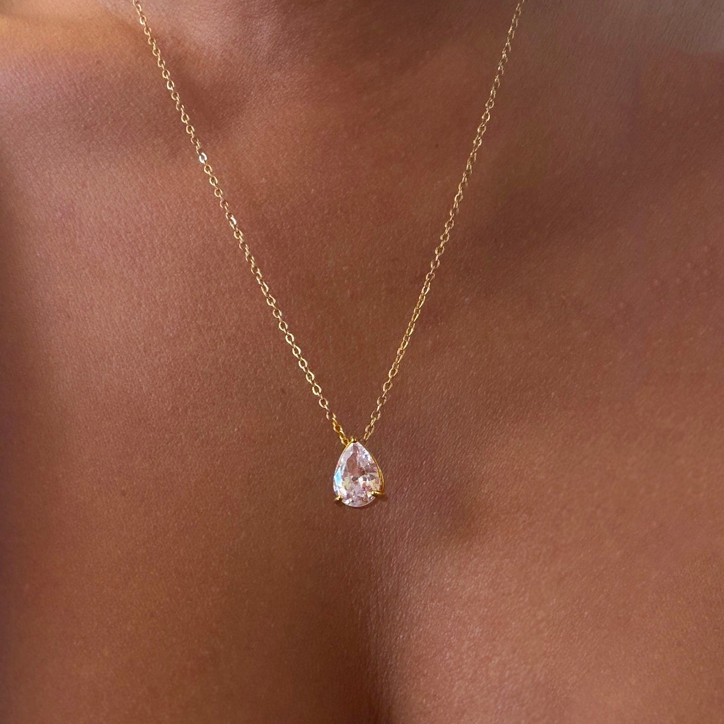 The Elara Necklace by LLUME Jewelry—a delicate 14K gold-plated chain adorned with a teardrop-shaped clear gemstone pendant—rests gracefully on medium brown skin.