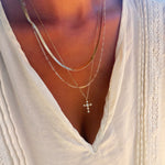 A person wearing the Ella Cross Favorites Set by LLUME Jewelry—a layered gold necklace featuring a striking paperclip chain and a cross pendant—over a cream-colored pleated blouse with lace trim and a deep V neckline.
