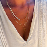 A person wearing the Ella Cross Favorites Set by LLUME Jewelry—a layered gold necklace featuring a striking paperclip chain and a cross pendant—over a cream-colored pleated blouse with lace trim and a deep V neckline.