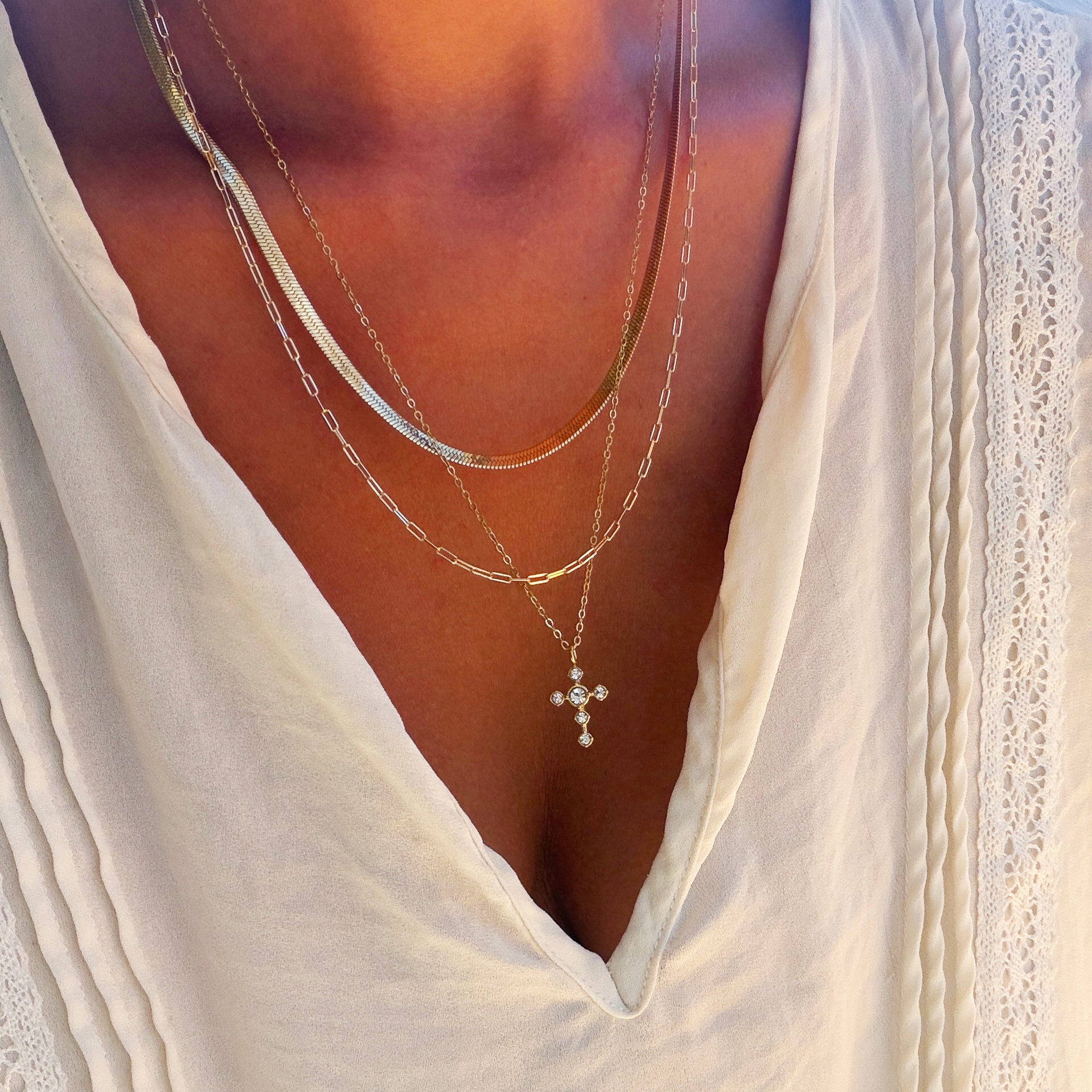 A person wearing the Ella Cross Favorites Set by LLUME Jewelry—a layered gold necklace featuring a striking paperclip chain and a cross pendant—over a cream-colored pleated blouse with lace trim and a deep V neckline.