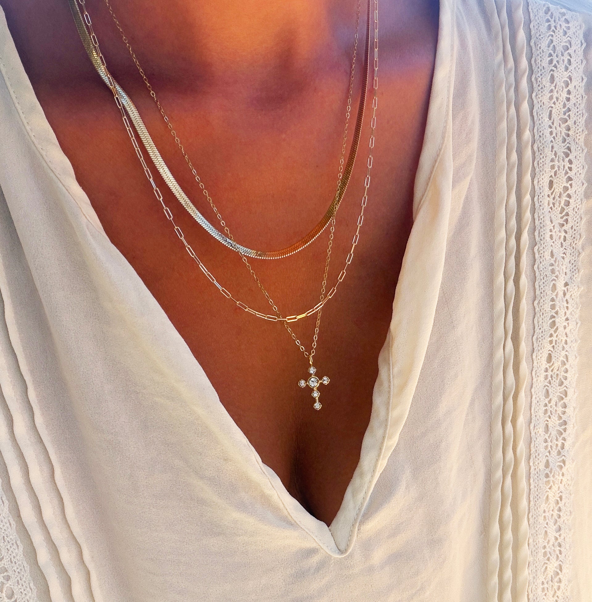 A person wearing the Ella Cross Favorites Set by LLUME Jewelry—a layered gold necklace featuring a striking paperclip chain and a cross pendant—over a cream-colored pleated blouse with lace trim and a deep V neckline.