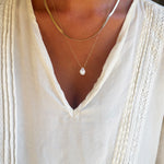 A person wearing a white blouse with lace detailing and pleats, accessorized with the Elara Classics Set from LLUME Jewelry, which features two layered necklaces: the Skinny Hailey chain and a gold solitaire teardrop pendant necklace adorned with a clear gemstone.