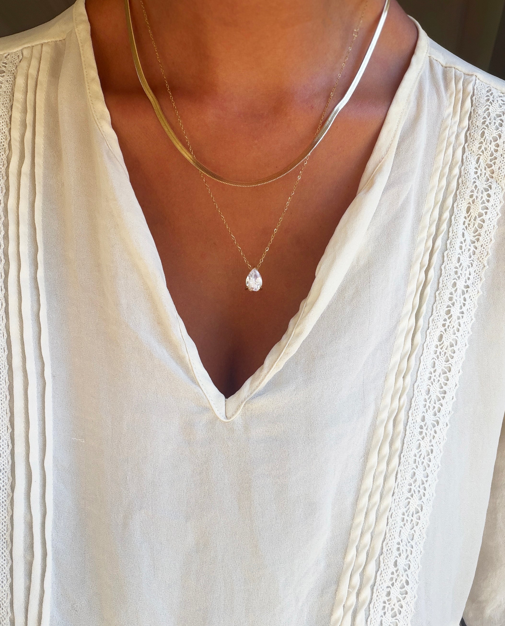 A person wearing a white blouse with lace detailing and pleats, accessorized with the Elara Classics Set from LLUME Jewelry, which features two layered necklaces: the Skinny Hailey chain and a gold solitaire teardrop pendant necklace adorned with a clear gemstone.