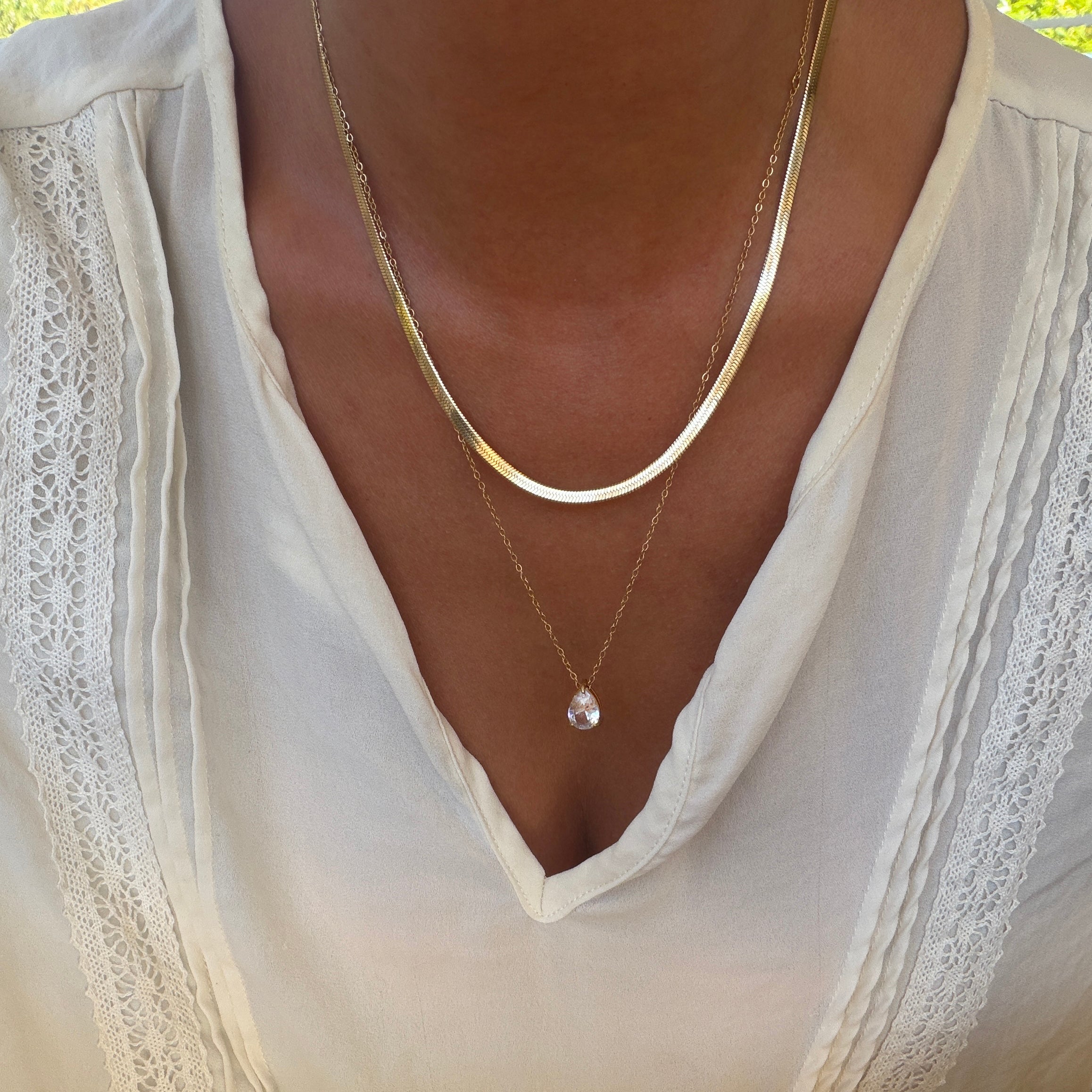 Adorned in a white blouse with lace accents, a person elegantly showcases the LLUME Jewelry Elara Classics Set—featuring two gold necklaces, including a skinny Hailey chain and a solitaire teardrop pendant—demonstrating the art of thoughtfully layered jewelry.