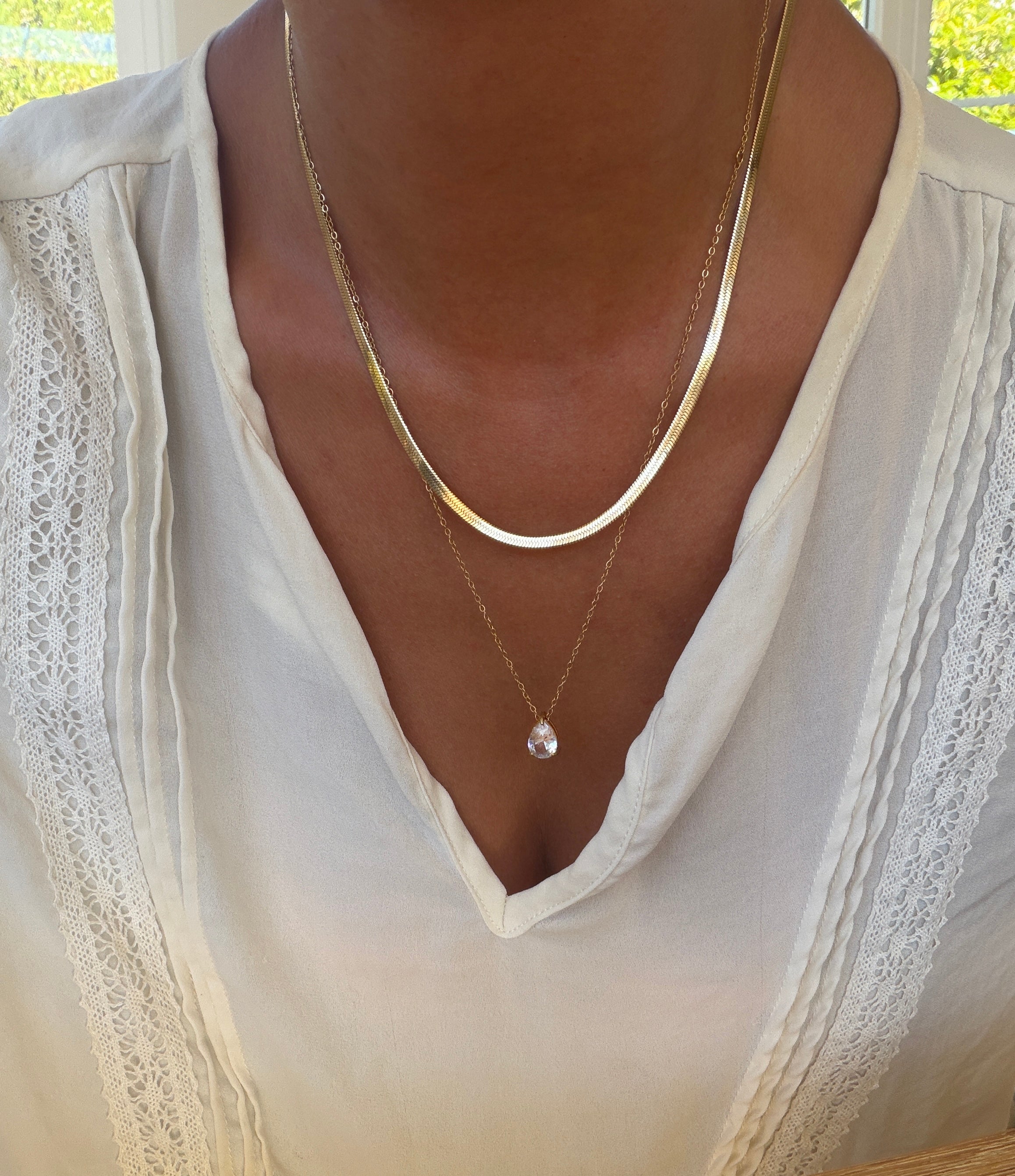Adorned in a white blouse with lace accents, a person elegantly showcases the LLUME Jewelry Elara Classics Set—featuring two gold necklaces, including a skinny Hailey chain and a solitaire teardrop pendant—demonstrating the art of thoughtfully layered jewelry.