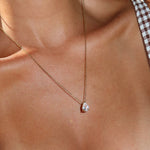 A close-up of a person’s neck wearing the LLUME Jewelry Elara Necklace, a 14K gold plated chain featuring a teardrop-shaped pendant. The person is dressed in a brown-and-white checkered top with thin straps.