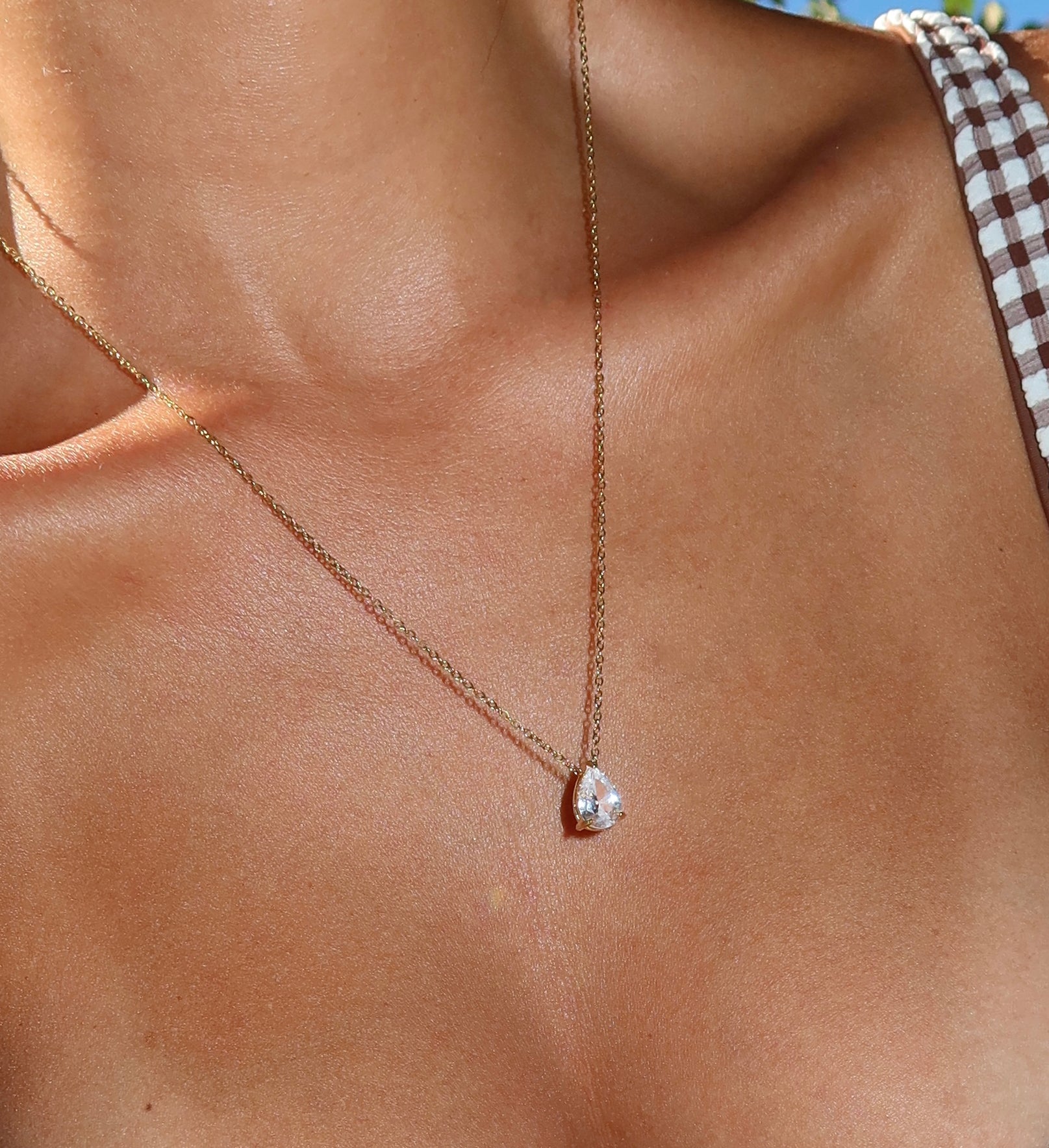 A close-up of a person’s neck wearing the LLUME Jewelry Elara Necklace, a 14K gold plated chain featuring a teardrop-shaped pendant. The person is dressed in a brown-and-white checkered top with thin straps.