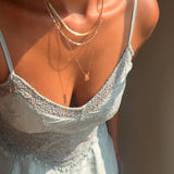 A woman in a light blue lace dress leans forward, bathed in warm sunlight, wearing the LLUME Jewelry Wild Honey Favorites Set—an elegant layered gold necklace set, with one chain featuring a rectangular pink pendant inspired by honeycomb motifs.