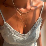 A person wearing a light blue lace dress and the LLUME Jewelry Wild Honey Favorites Set—a stunning trio of layered gold necklaces, one adorned with a rectangular pendant—illuminated by warm sunlight.