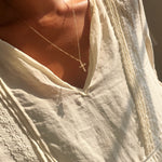 A person wearing the Ella Cross Necklace by LLUME Jewelry—a delicate, gold-plated necklace with a small cross pendant—paired with a cream-colored, lace-trimmed blouse, photographed from the shoulders to upper chest in warm sunlight.