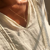A person wearing the Ella Cross Necklace by LLUME Jewelry—a delicate, gold-plated necklace with a small cross pendant—paired with a cream-colored, lace-trimmed blouse, photographed from the shoulders to upper chest in warm sunlight.