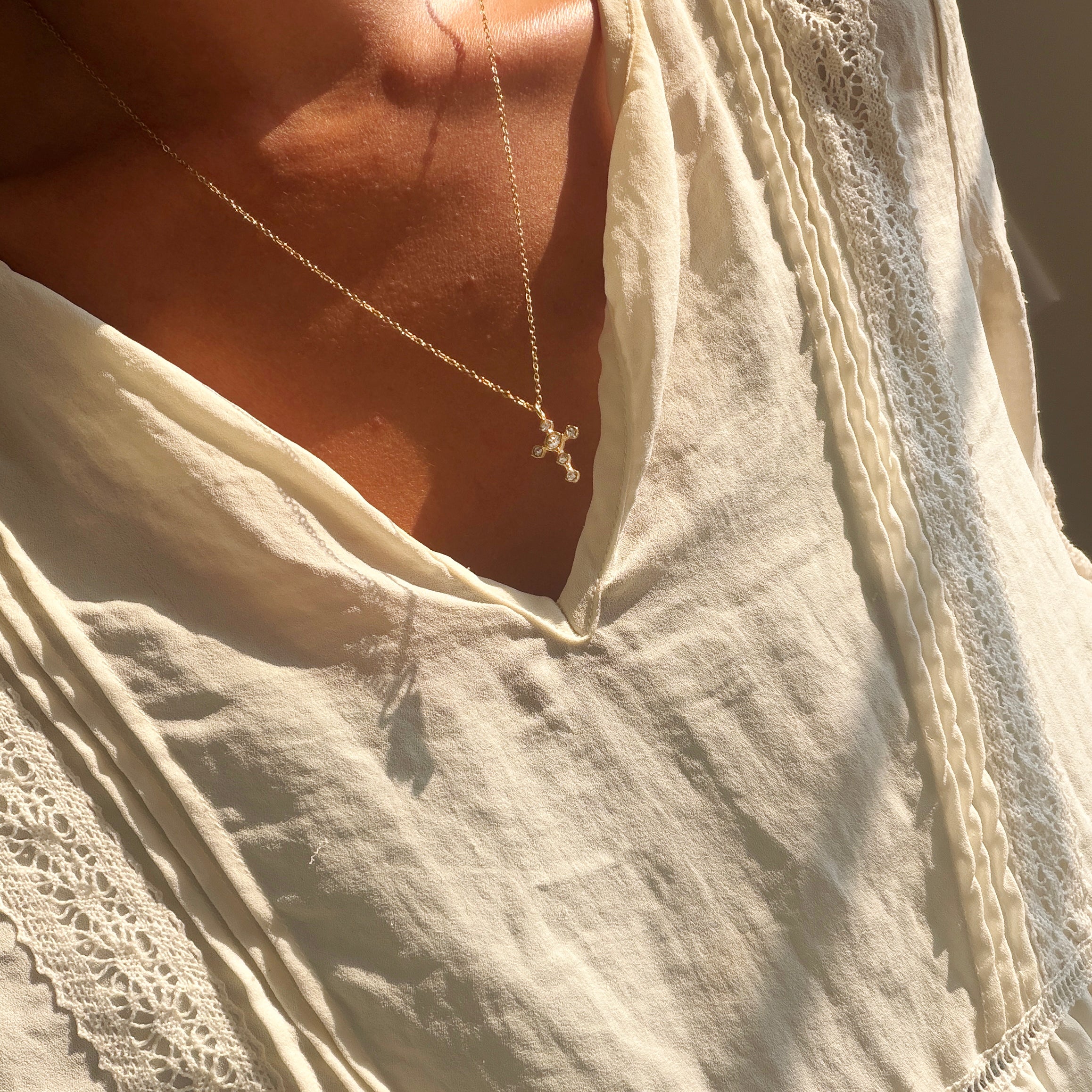 A person wearing the Ella Cross Necklace by LLUME Jewelry—a delicate, gold-plated necklace with a small cross pendant—paired with a cream-colored, lace-trimmed blouse, photographed from the shoulders to upper chest in warm sunlight.