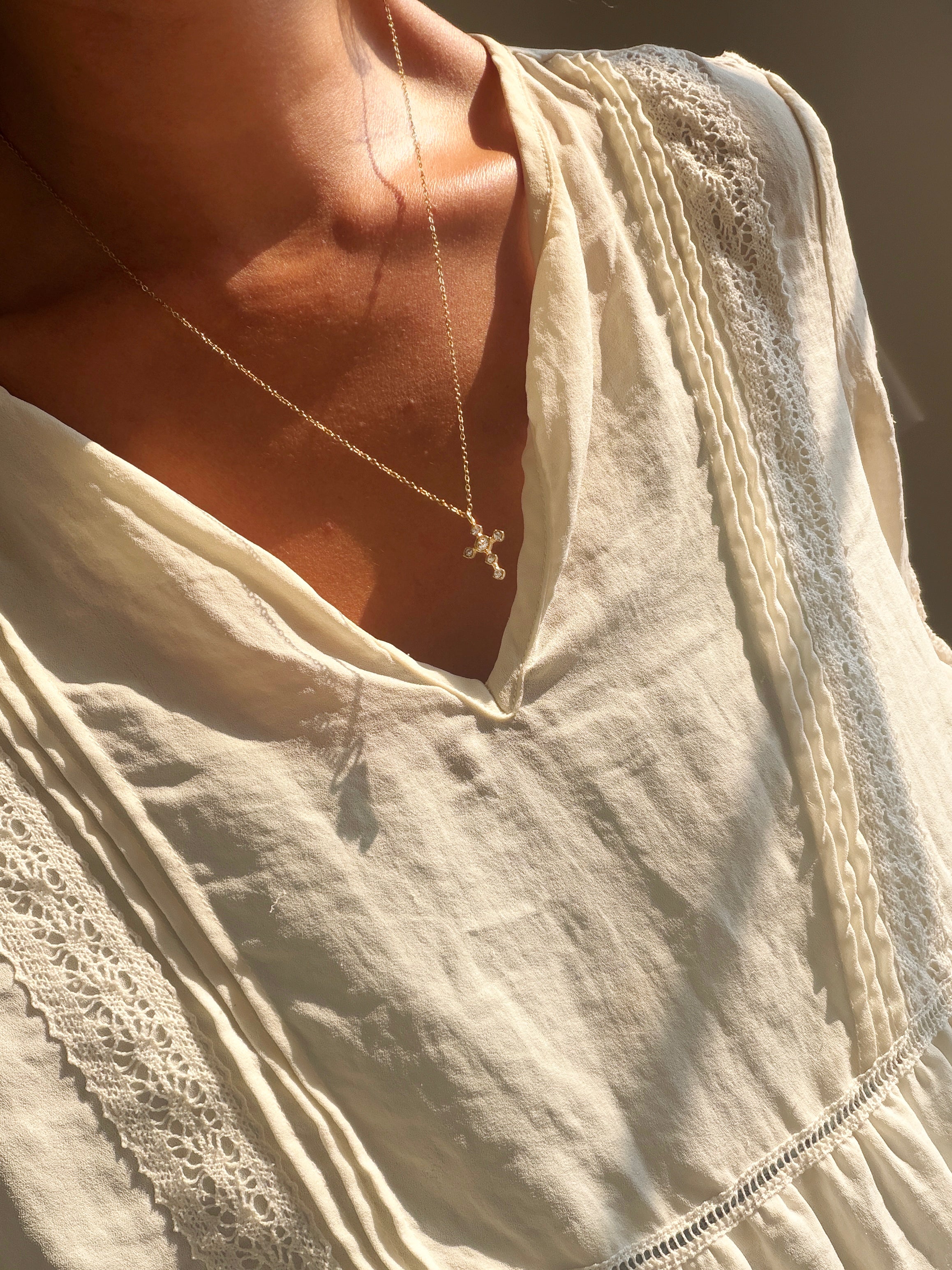 A person wearing the Ella Cross Necklace by LLUME Jewelry—a delicate, gold-plated necklace with a small cross pendant—paired with a cream-colored, lace-trimmed blouse, photographed from the shoulders to upper chest in warm sunlight.