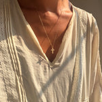 A person wearing a cream-colored, textured blouse and LLUME Jewelry’s Ella Cross Necklace—gold plated and waterproof—stands with sunlight illuminating their skin and clothing, showcasing both style and durability.