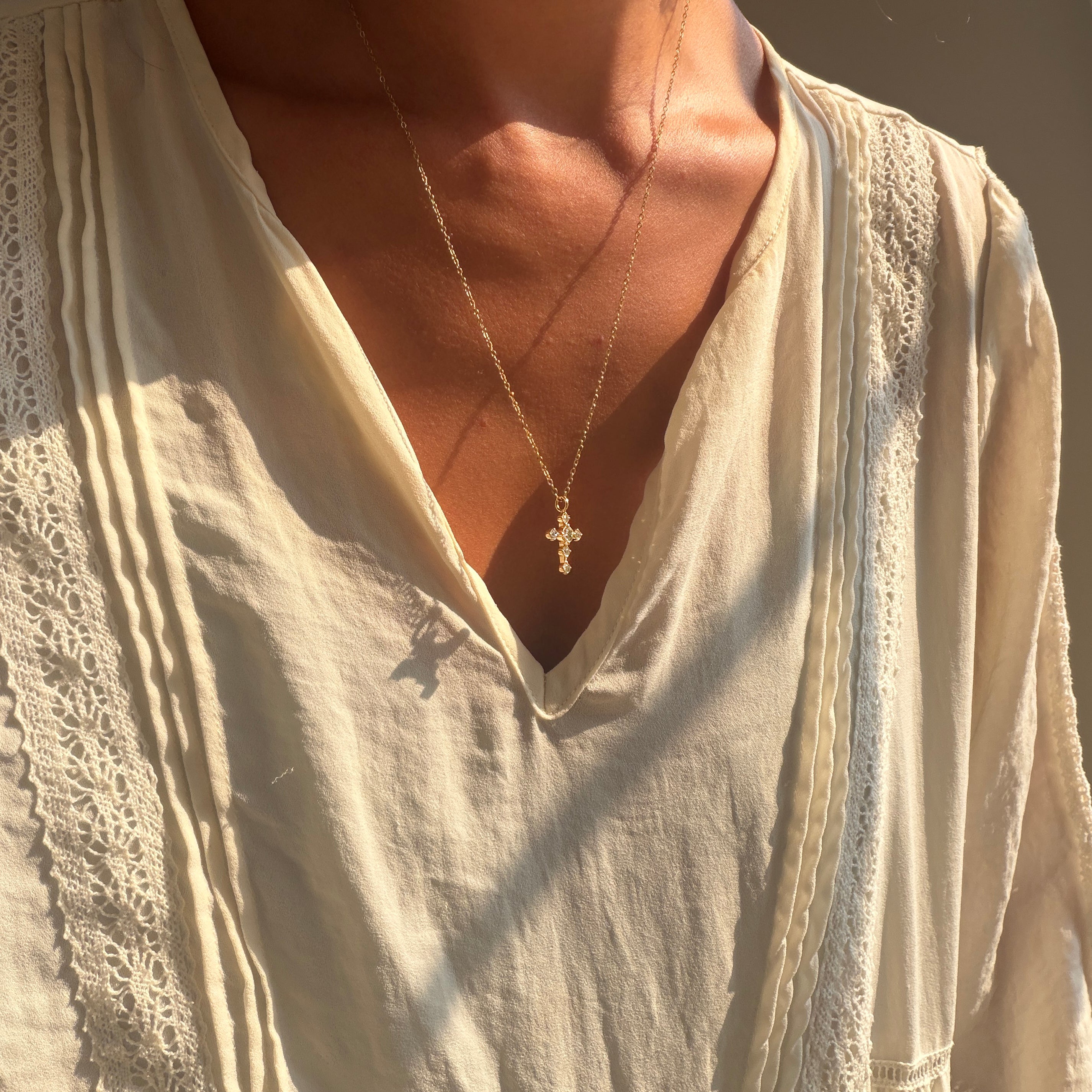 A person wearing a cream-colored, textured blouse and LLUME Jewelry’s Ella Cross Necklace—gold plated and waterproof—stands with sunlight illuminating their skin and clothing, showcasing both style and durability.