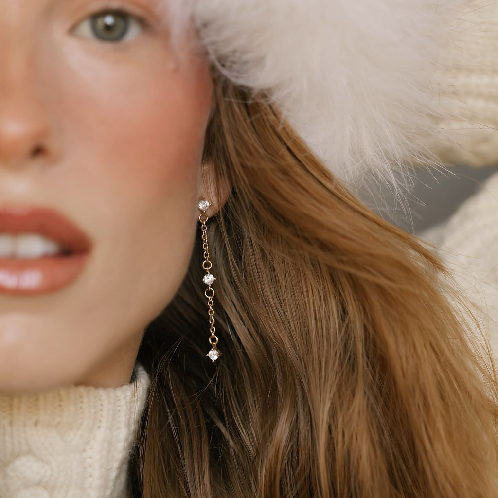 A close-up of a woman with wavy brown hair wearing a white fuzzy hat, white knit sweater, and LLUME Jewelry’s Reverie Earrings—elegant drops that shimmer. She has light skin and natural makeup with rosy cheeks.